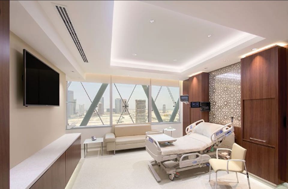 Interior of a premium fit-out with full-height glazing overlooking a city skyline, a recessed cove-lit ceiling, warm walnut timber joinery, a wall-mounted screen, large-format light stone flooring, and a feature mosaic accent panel.