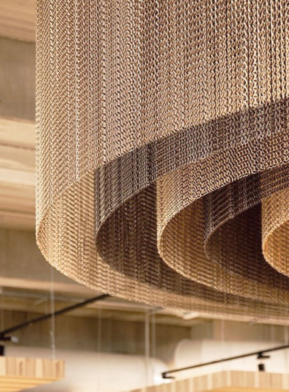 Suspended bronze chainmail-mesh ceiling feature with sweeping scalloped curves above a retail concourse, set against light timber-slatted ceilings and walls, exposed concrete soffits and black track lighting.